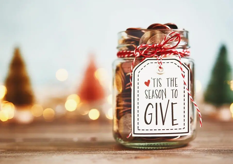 Glass jar filled with coins and a holiday label, set among colorful trees and lights, promoting charitable giving advice.
