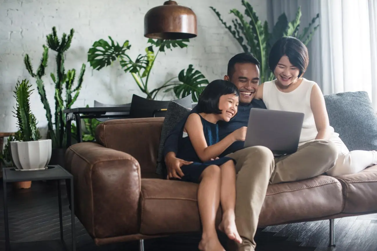 Father, mother, and daughter on a sofa with a laptop, discussing financial literacy in a bright, modern room.