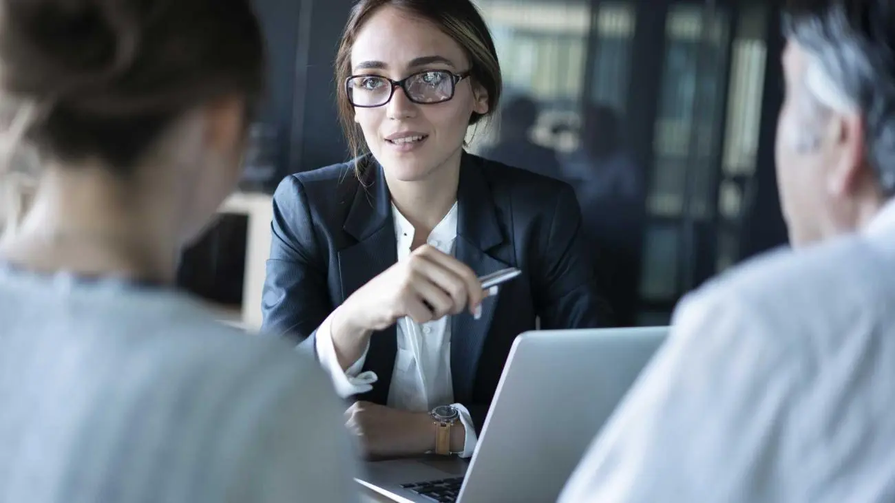 Young woman in a formal suit discussing financial planning with a colleague, laptop and pen on table.