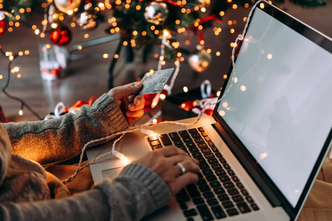 Person with a credit card shopping online by a decorated Christmas tree in a cozy setting, by Statera Financial Planners.