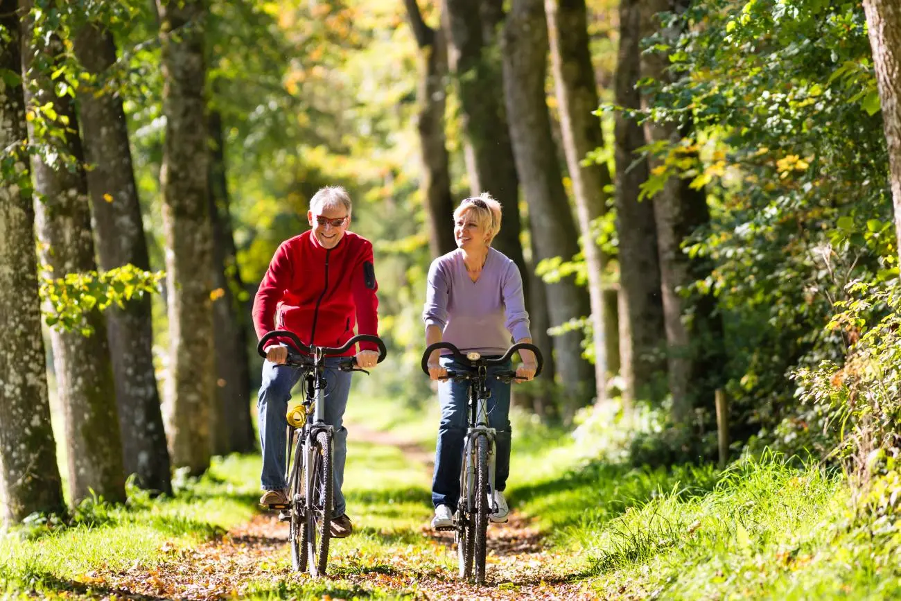 Two middle-aged cyclists wearing helmets ride bikes along a sunlit path with autumn foliage, representing active lifestyles.