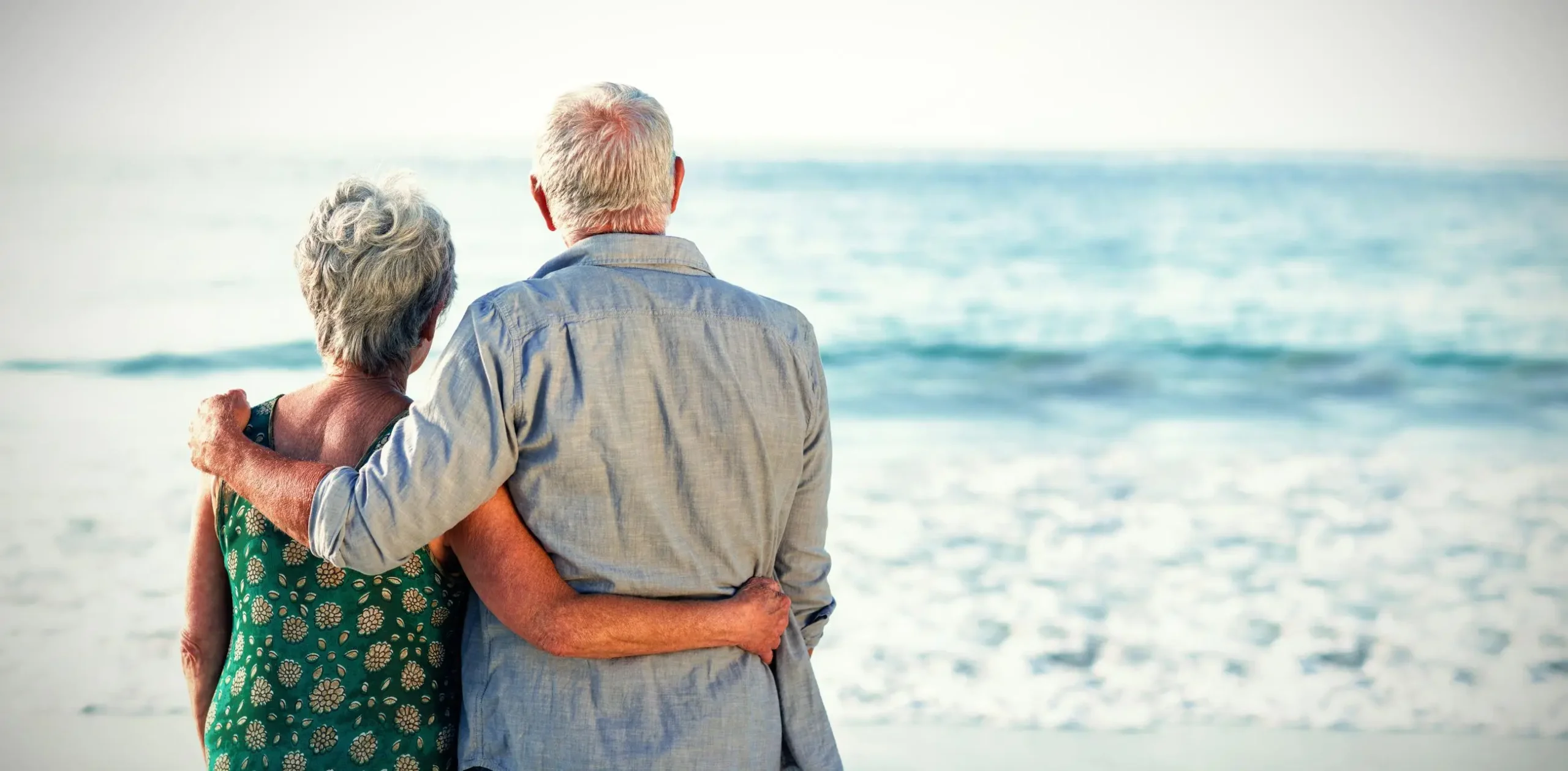 Elderly couple embracing by the ocean, representing companionship as they plan retirement with Statera Financial Planners.